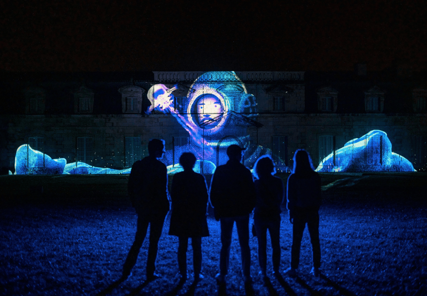 Cinq personnes se tiennent face à la Corderie Royale et admirent une illumination avec un homme en scaphandre